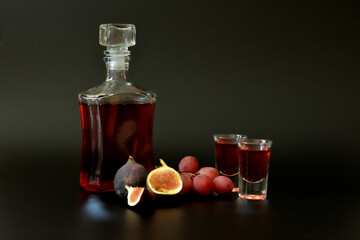 Fig-grape liqueur in a bottle and two glasses on a black background, next to ripe fruits.
