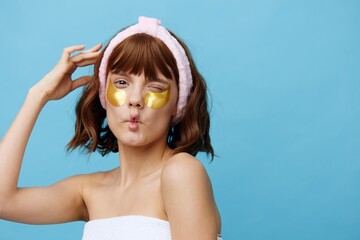 Horizontal photo, a woman with ideal smooth skin on a blue background with luxurious dark hair and a cute pink rim put on golden eye patches and is very surprised at what is happening around