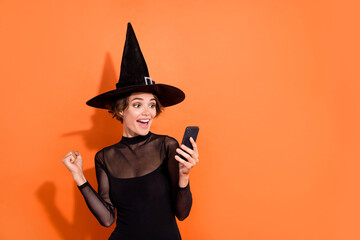 Photo of delighted excited sorcerer girl raise fist success use telephone instagram twitter facebook telegram isolated on orange color background