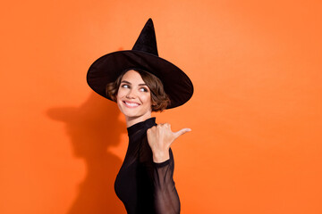 Photo of cute cheerful young lady wear black witch dress looking pointing back empty space isolated orange color background