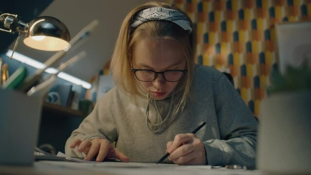 Focused illustrator draws sketches for her project. Young female artist with glasses dressed in a grey hoodie and sits on her work desk. Makes pencil sketches. Story telling concept.
