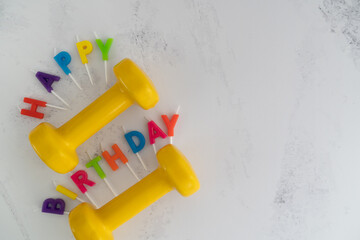 Colorful Happy Birthday cake candles letters and yellow dumbbells. Exercise equipment as a gift idea, flat lay composition. Gym workout training concept with copy space. Top view party invitation.