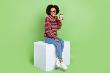 Full body photo of nice young curly hairdo lady sit on podium drink wear eyewear sweater jeans footwear isolated on green background
