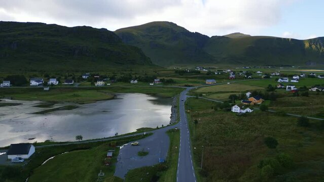 Flying downwards towards street road in Lofoten, Norway with surrounding fairytale mountain and green grass landscape with tiny houses along the road