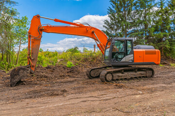 Orange new crawler excavator stands against backdrop of green trees and blue cloudy sky.
