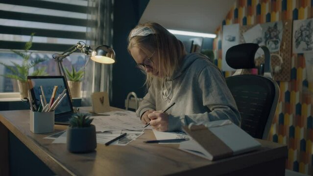 A blonde woman working on a storyboard in a home based design studio. Focused woman dressed in a hoodie drawing pencil sketches. Making storyboard as a roadmap for the video.
