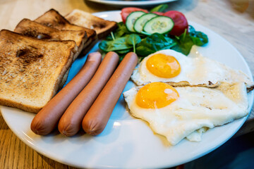 Delicious breakfast prepared with three sausages, fried eggs and four pieces of toasted bread served in white plate with fresh spinach, tomato and cucumber slices.