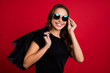 Photo of funny cool young lady dressed black t-shirt arm dark glasses smiling holding bargains isolated red color background