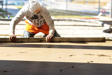 Close up of man builder placing screed rail on the floor covered with sand-cement mix at construction site. Male worker leveling surface with straight edge while screeding floor. Blurred background.