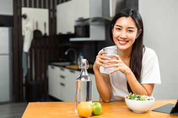 Young female drinking water in glass for refreshing for diet and wellness on table. Beautiful woman happily drink fresh water before breakfast in kitchen in the morning. Healthy food concept.