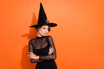 Photo of shiny thoughtful young lady wear black witch dress cap arm crossed looking empty space isolated orange color background