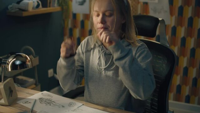 Young creative designer walks towards her work desk in a home based studio. The woman sits on the chair, puts on her glasses and starts sketching with a pencil.