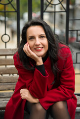 brunette woman in red coat with red lips sitting on the bench in autumn Kyiv city