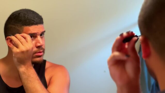 Gay mixed race man looking into mirror adding eyebrow makeup in bathroom, close up LGBTQ