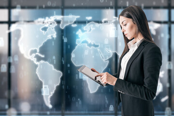 Attractive young european businesswoman using tablet and standing in interior with creative map hologram on blurry window with city view. Global business, success and CEO concept.