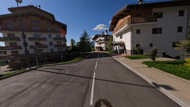 Aerial view white speed sports car riding asphalt road at suburb alpine resort street infrastructure mountain sunny snowy peak. FPV drone shot traffic roadway at town landmark village house exterior