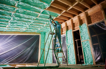 Male builder insulating wooden frame house. Man worker spraying polyurethane foam inside of future...