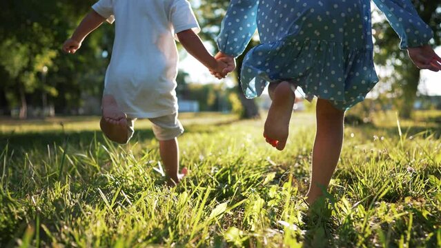 Happy family.Toddlers run barefoot on the green grass in park at sunset.Active running in nature.Summer picnic healthy lifestyle.Legs of children in park on grass.Brother and sister outdoors in summer