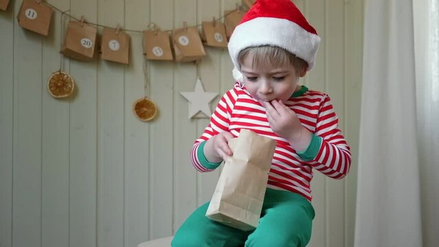 Boy in pajamas, Santa cap eating sweet candy on paper advent calendar with presents background. Little happy child with gift of handmade Christmas calendar on wall. Celebrating at home Xmas tradition.