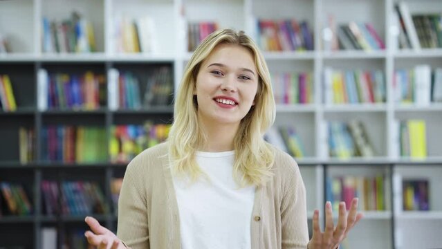 Young cheerful female blogger influencer talking to camera, making video chat or conference call. Happy girl giving interview. Student recording vlog in library about her college