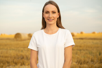 A young woman in a white T-shirt against the background of haystacks looks into the camera, a beautiful romantic girl outdoors in a field at sunset.