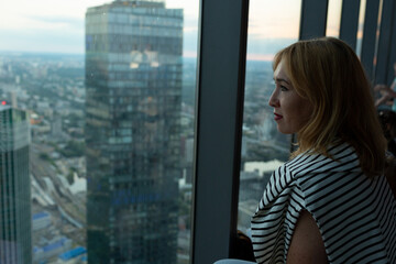 A woman stands at a panoramic window in a high-rise building