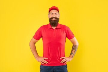 Happy bearded man in red cap and tshirt standing with tattooed arms akimbo yellow background, casual