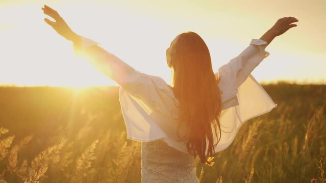 Woman half-turned with flowing hair on a blooming field looks at the sunset. A girl with long hair fluttering in the wind raises her hands up. 
