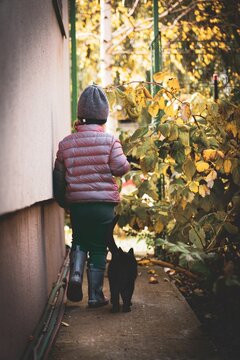 Vertical Shot Of A Kid Walking With A Black Cat In The Backyard