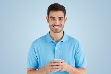 Young man standing on blue background, holding smartphone, looking at camera and smiling nicely