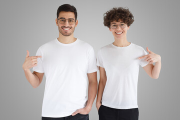 Smiling couple pointing at blank white t-shirts with index fingers, copy space for your advertising