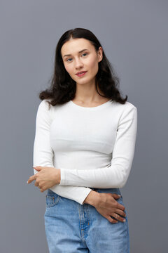 a gentle woman stands on a dark background in a tight white T-shirt, hugs herself a little with her hands and looks sweetly to the side