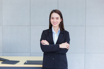 Asian professional working woman who has long hair wears black formal suit with blue shirt while she arm crossing and confident smiles in office room.