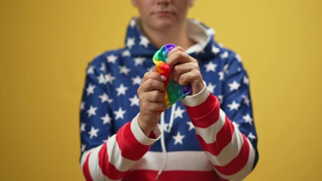 Nervous anxious millennial lady sqeezing colorful sensory toy throwing away standing at yellow background. Young Caucasian stressed woman in US-flag colored hoodie with fidget toy. Stress management
