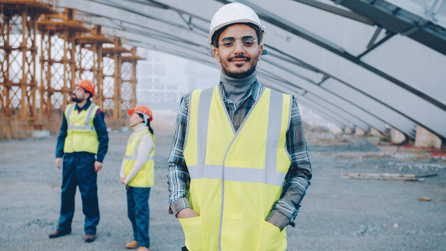 portrait of Arab man wearing safety helmet standing in construction site smiling looking at camera while people in uniform working in background