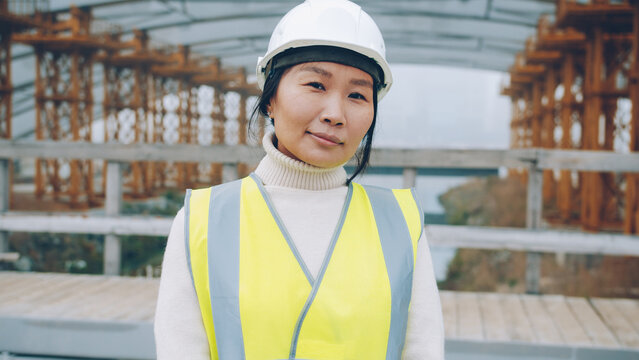 portrait of female builder standing outside construction area wearing safety helmet. People and technical profession concept.