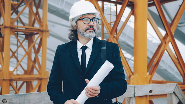 Portrait of serious mature man architect standing in construction site with blueprint looking around wearing white helmet and suit. People and occupation concept.