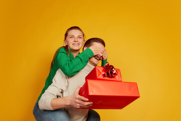Portrait of loving couple, man and woman celebrating holidays, making surprise with presents isolated over yellow background