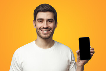 Young smiling man showing blank screen of smart phone in hand standing on yellow background