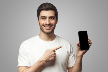 Young smiling man holding blank phone with empty black screen, pionting at display with finger