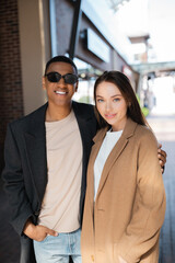 stylish african american man in sunglasses holding hand in pocket and hugging girlfriend in trendy coat.