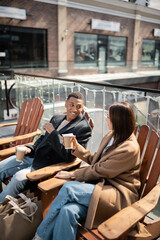 cheerful multiethnic couple in coats sitting with coffee to go near shopping bags and blurred mall.
