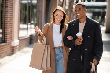 interracial couple in trendy coats smiling while walking in city with shopping bags and coffee to...