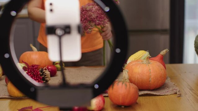 female blogger recording video using smartphone thanksgiving holiday decoration fall crop autumn ripe vegetables on kitchen table. woman live streaming mobile phone tripod circular led lamp pumpkins
