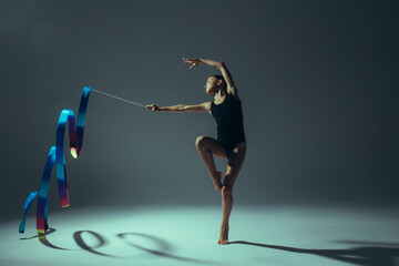 Gymnast child girl lit by light performs gymnastic exercises with ribbon in her hand.