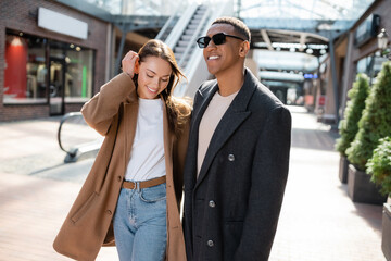 stylish african american man in sunglasses smiling near girlfriend fixing hair on street.