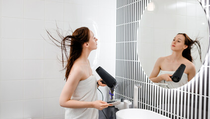 Young woman drying and styling hair with hair dryer, making hairstyle in modern bathroom interior. Beauty routine after morning shower