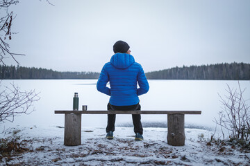 Man in a blue jacket with thermos sits on wooden bench on the shore of a frozen lake. Man in focus,...