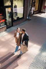 high angle view of interracial couple in trendy coats walking with shopping bags and coffee to go...