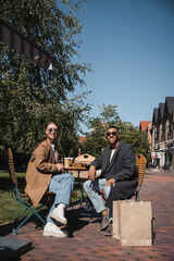 happy multiethnic couple in coats and sunglasses sitting near coffee to go and shopping bags in street cafe.